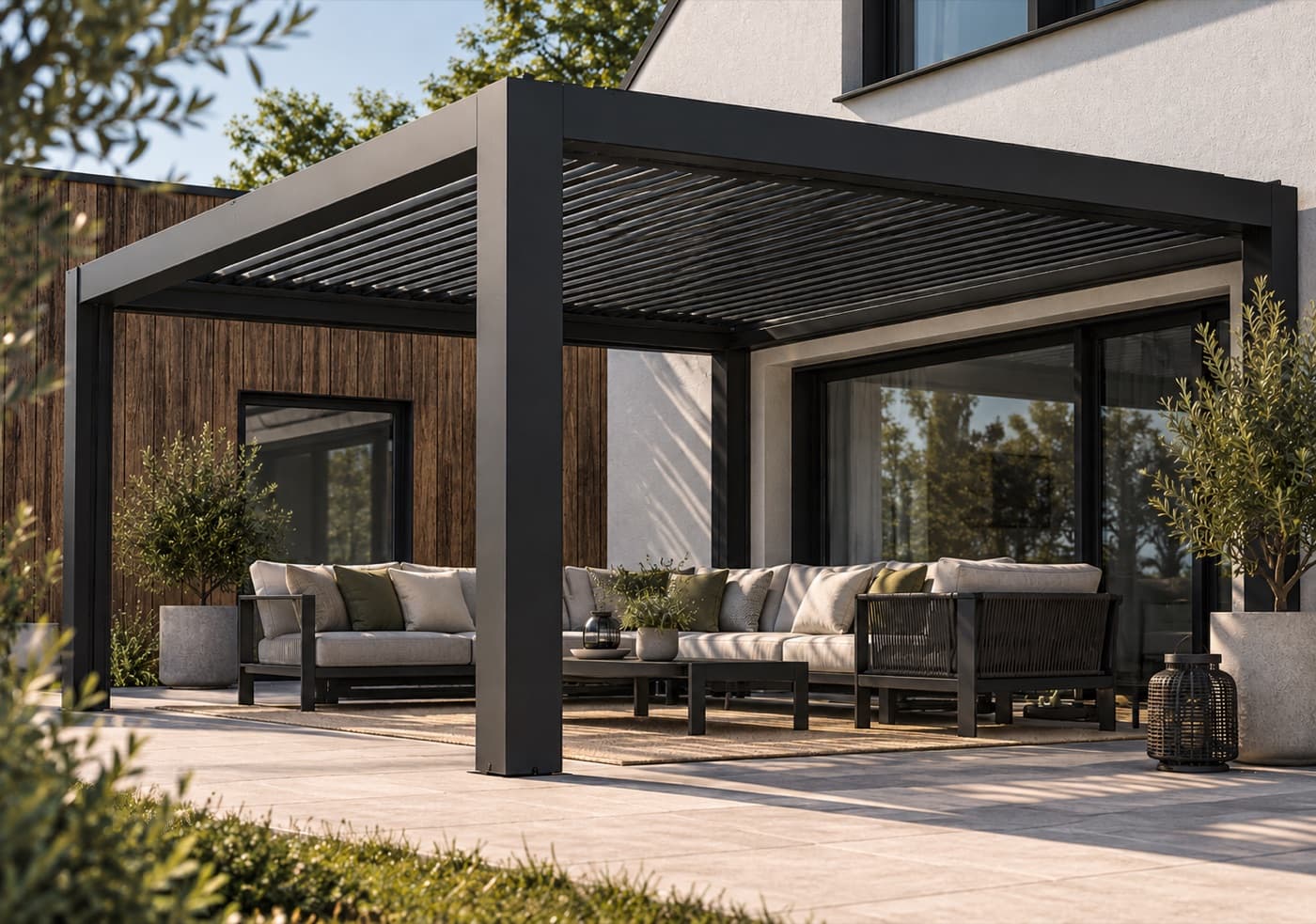 Antracitgrå bioklimatisk pergola med öppet lamelltak mot en ljus husvägg, med loungegrupp under taket